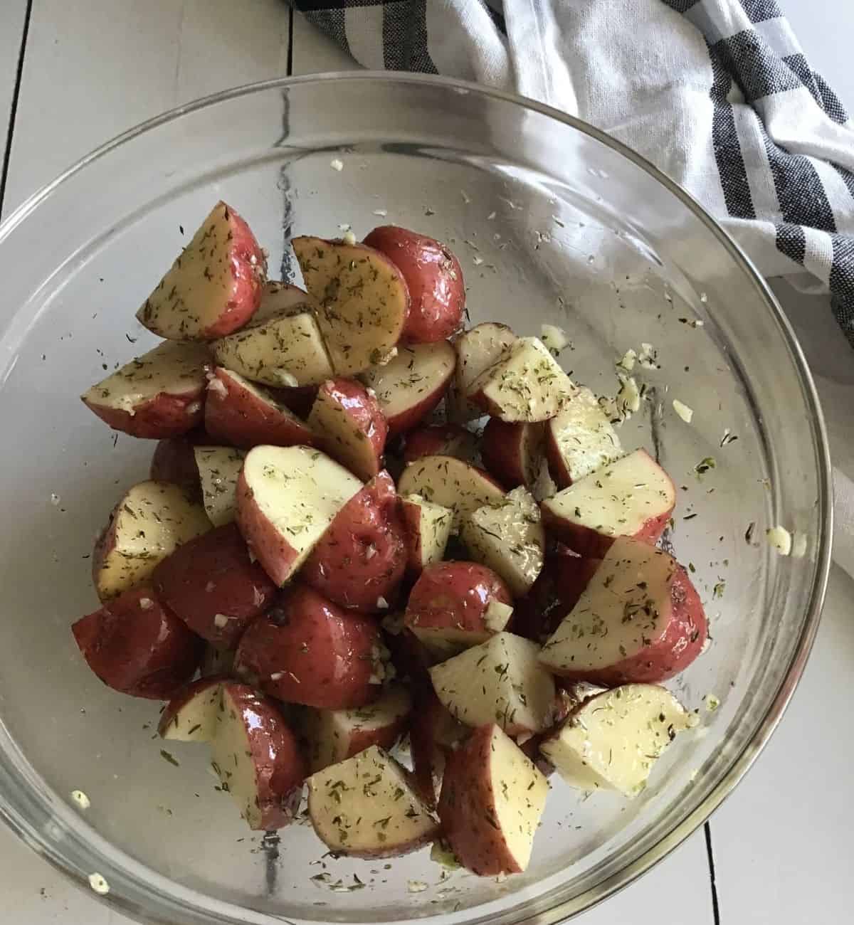 A glass bowl with cut red potatoes covered with olive oil, herbs, and garlic. 