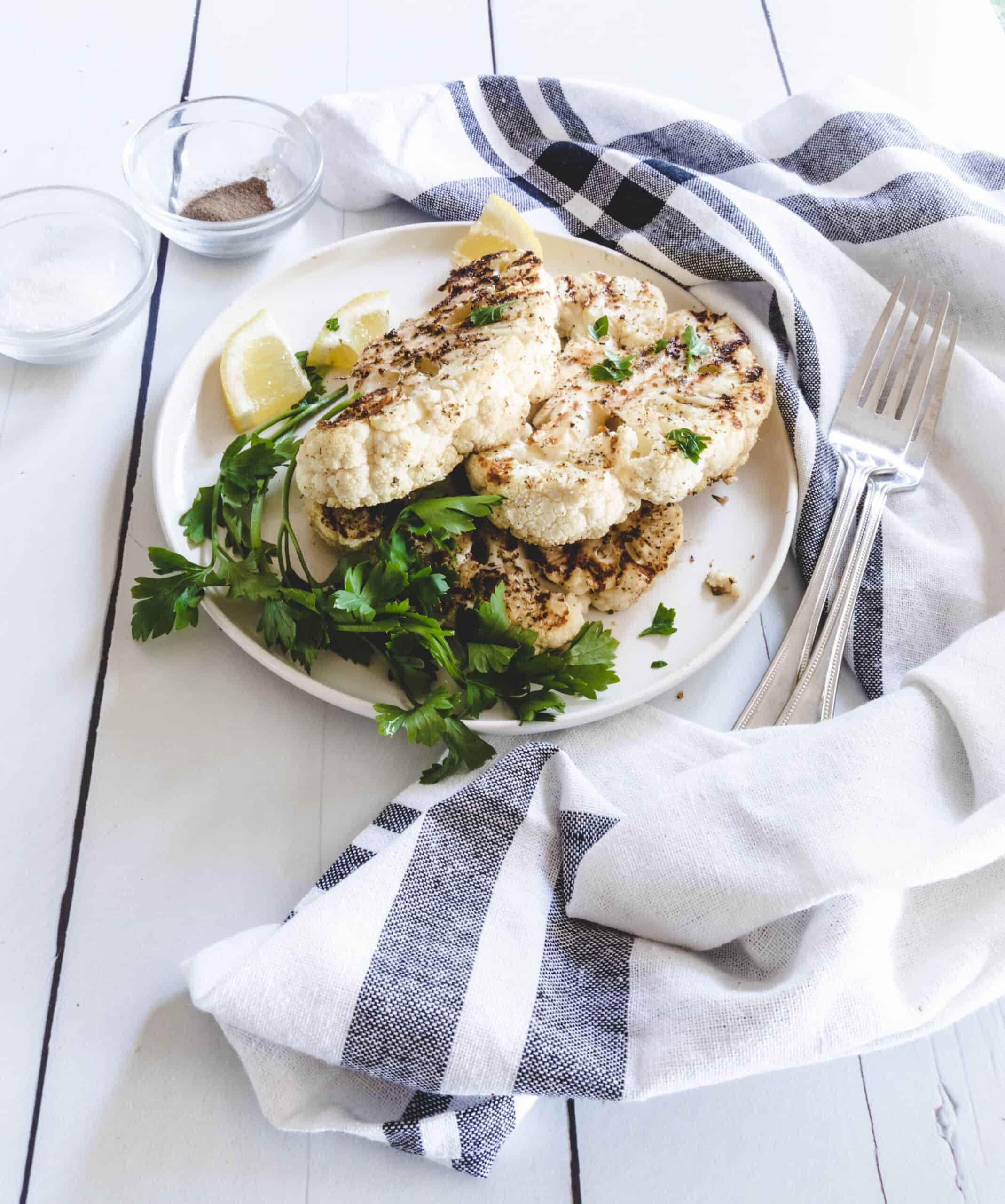 A white plate with a blue and white linen on the side wth thick sliced cauliflower on the plate.