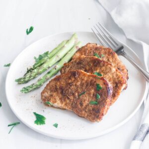 A white round plate with 3 cooked boneless pork chops with asparagus on the side.