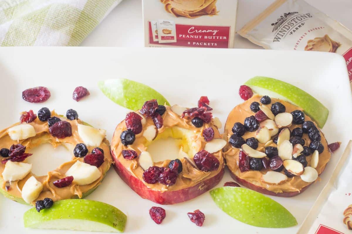 A white rectangle plate with three apple slices topped with peanut butter, dried fruit, and almond slices.