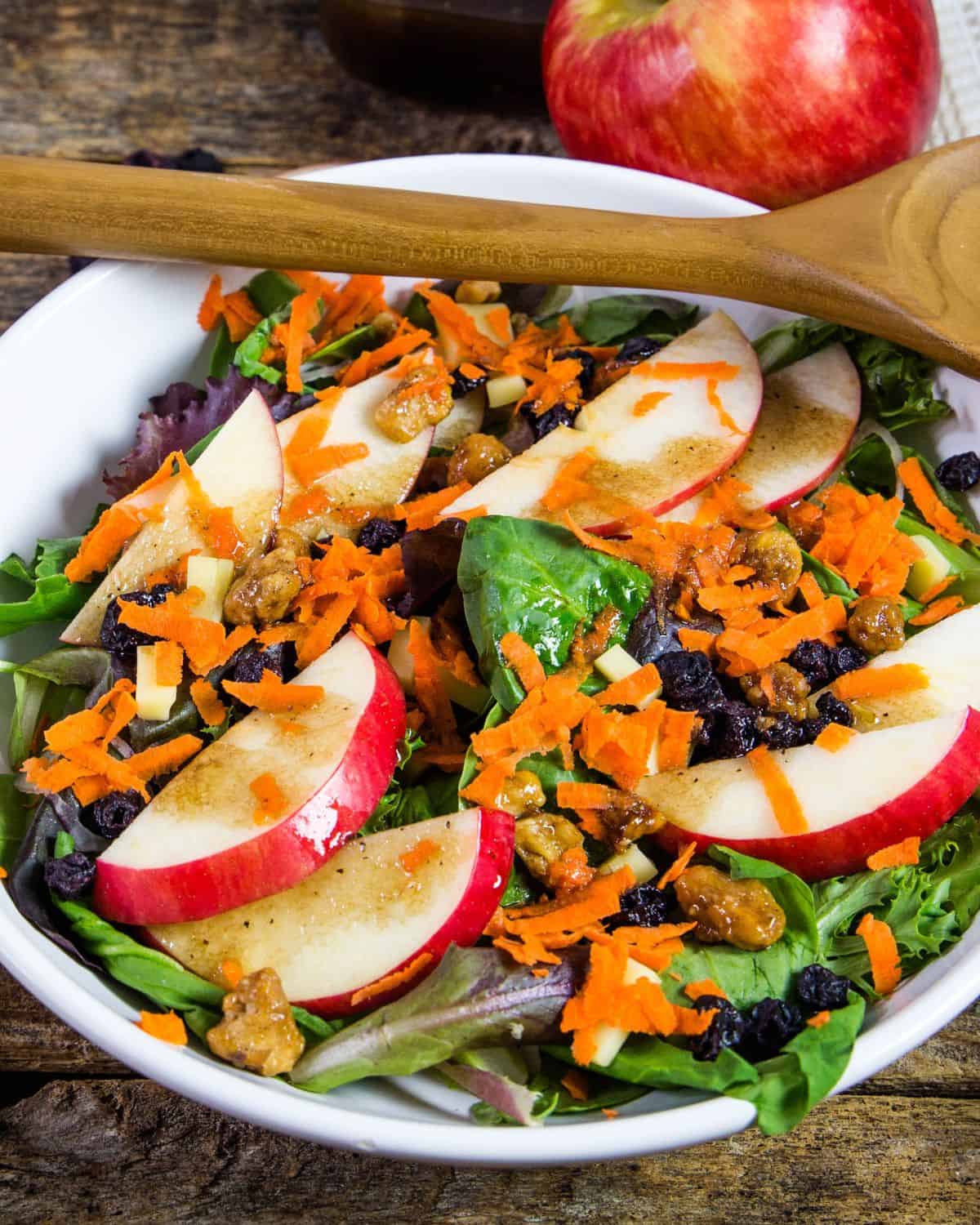 A white bowl filled with mesclun greens, apples, and shredded carrots with a vinaigrette on top.