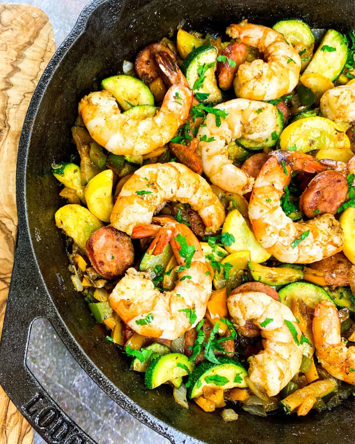 A cast iron pan with sauteed veggies, sausage and large cooked shrimp with cajun seasoning.