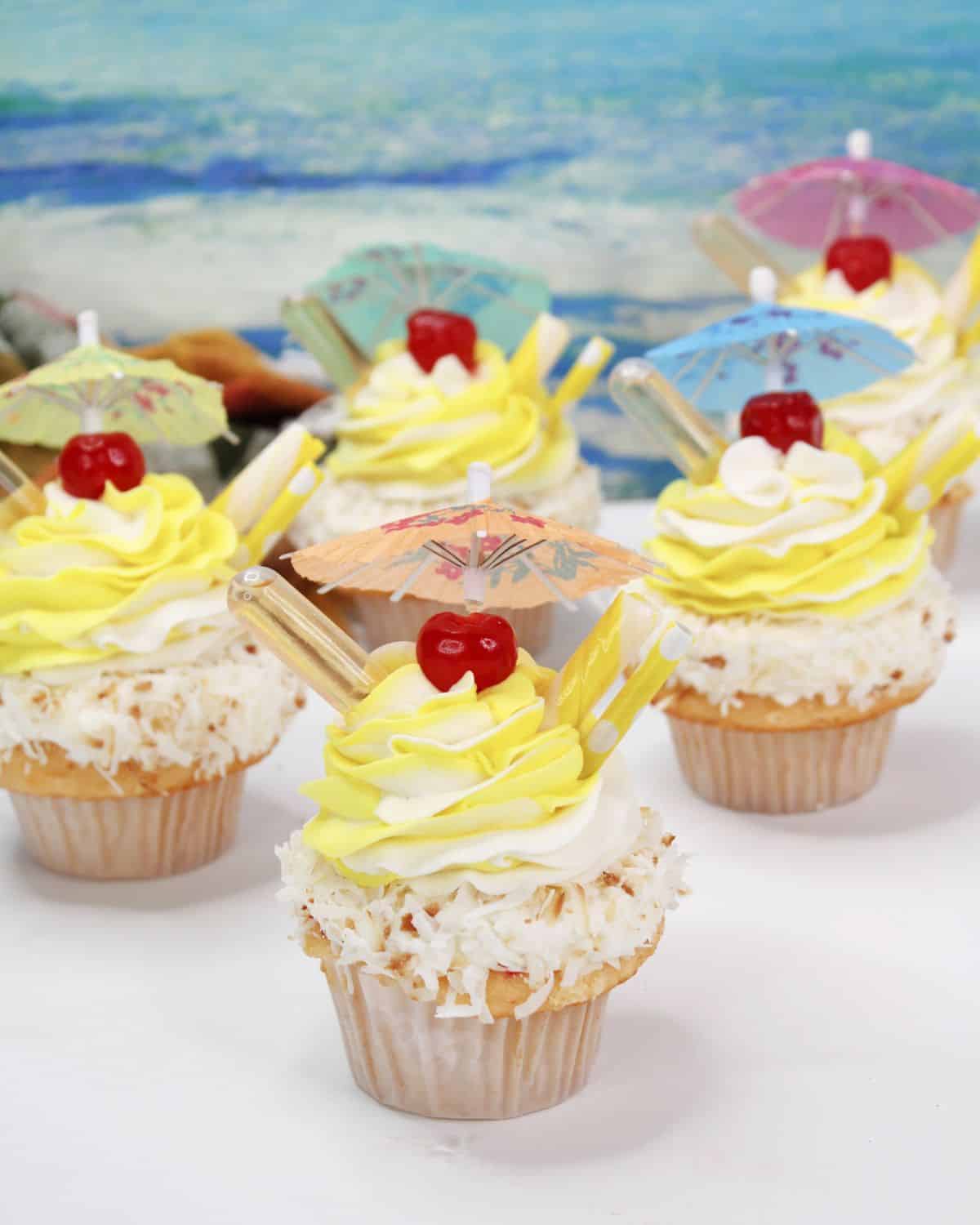 Four pina colada cupcakes with shredded coconut, topped with yellow frosting, cherry, and a tiki drink umbrella.