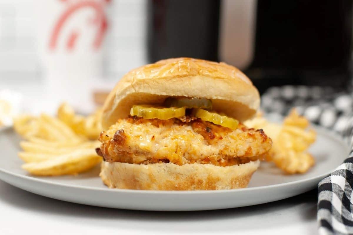 A side view of a baked chicken sandwich topped with pickles on a bun with waffles fries on a grey plate. 