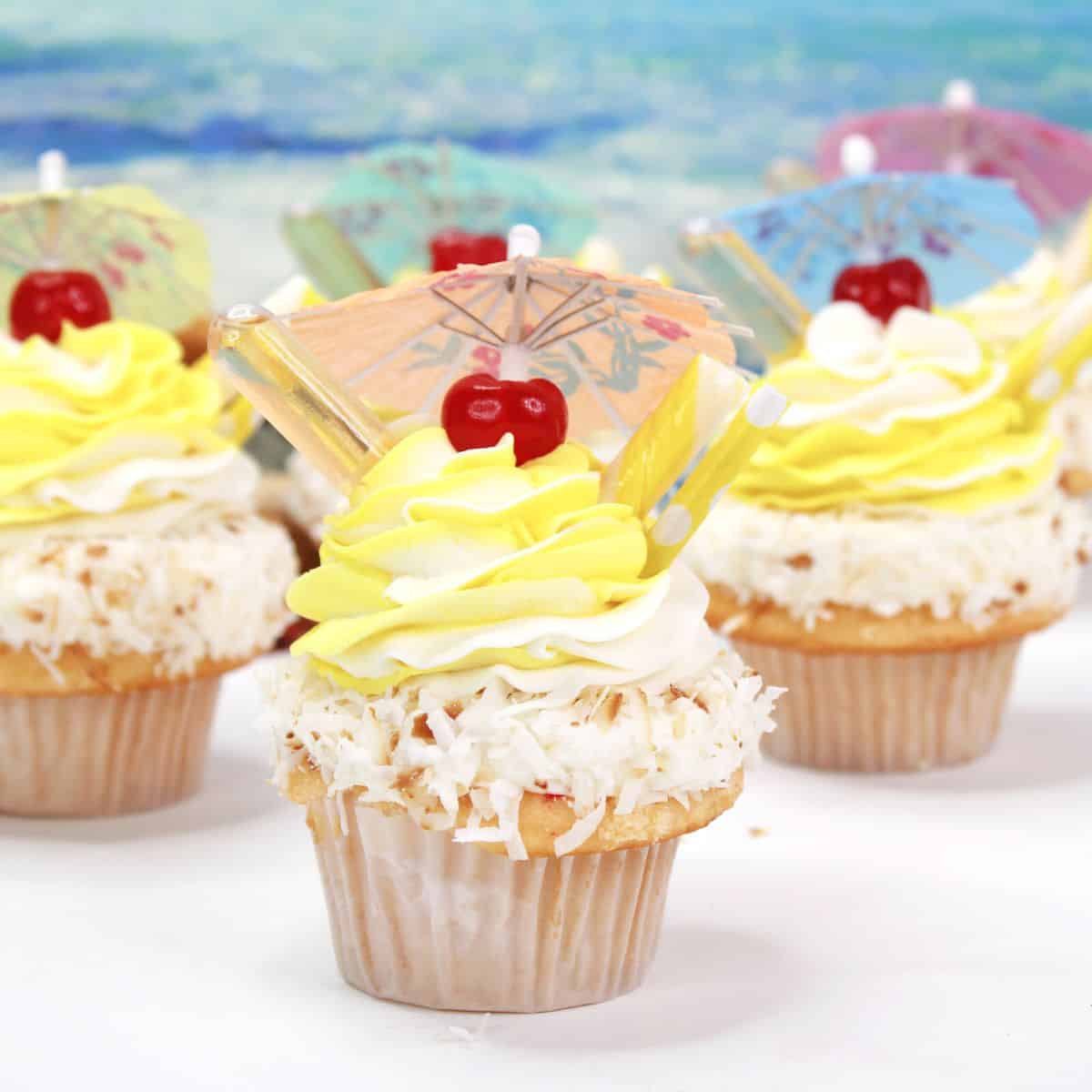 Three pina colada cupcakes with coconut and yellow frosting topped with a cherry and an umbrella.