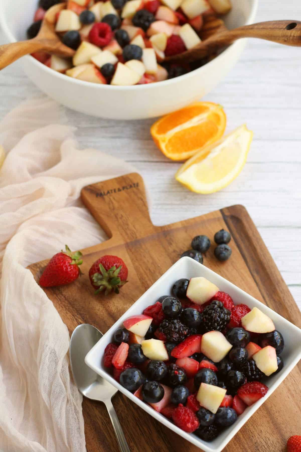 An overhead view of a square bowl filled with fruit on a wooden board with a peeled orange behind the cutting board.