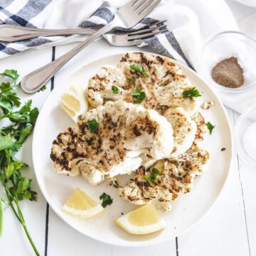 Grilled cauliflower steaks on a white plate with lemon wedges on the plate.