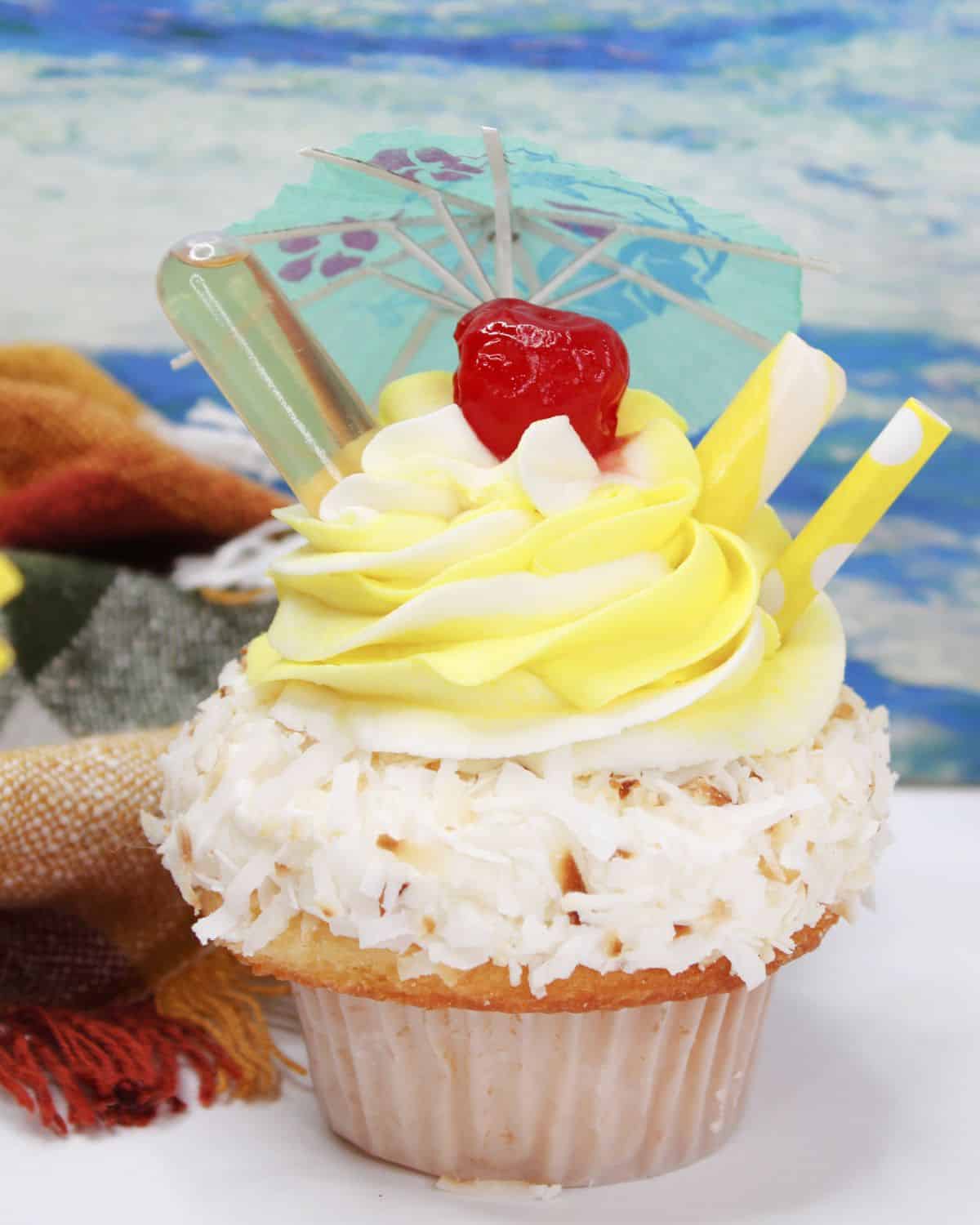 A cupcake with coconut flakes and yellow buttercream frosting with straws, a cherry, and an umbrella on the top