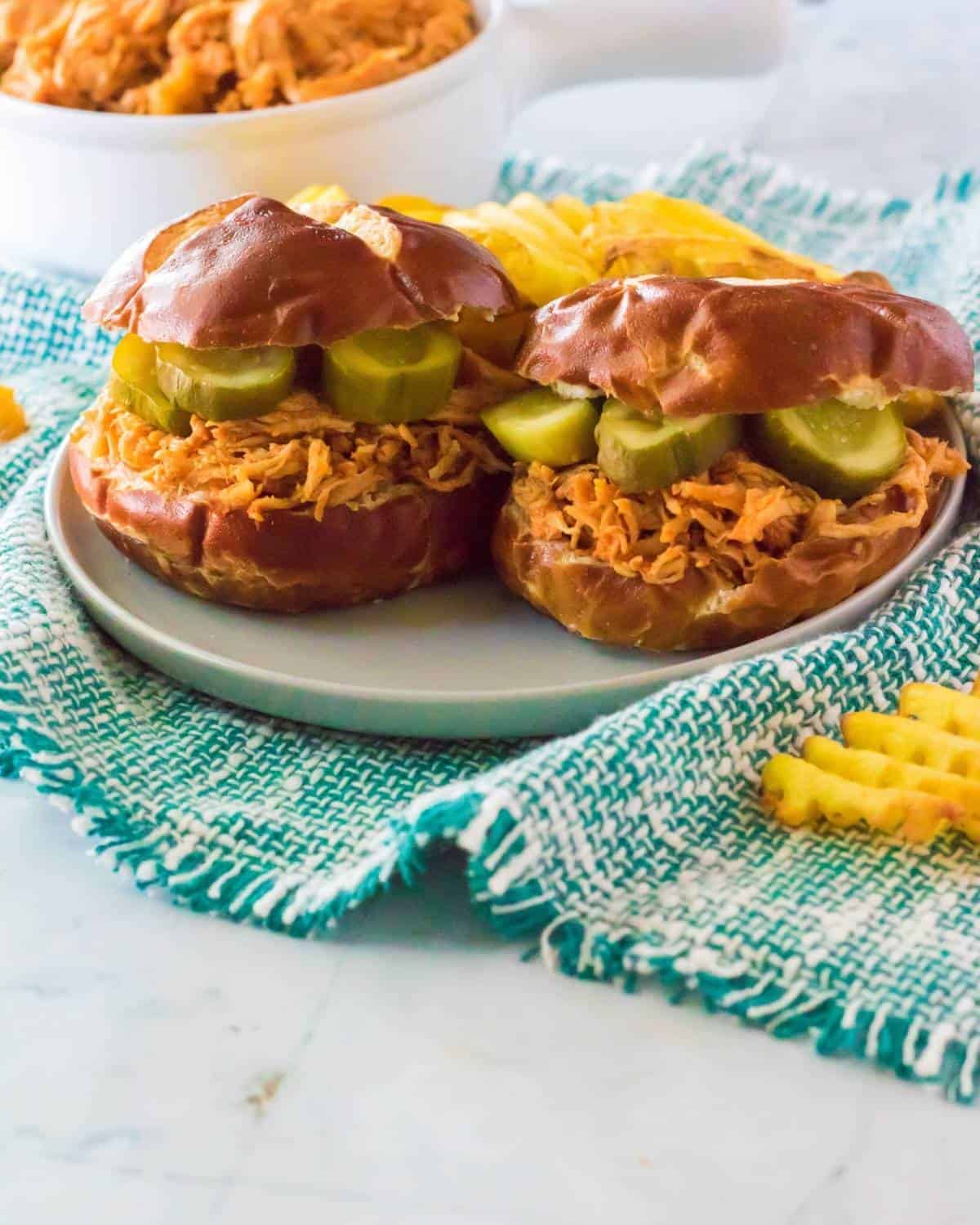 A shredded chicken sandwich on a pretzel bun topped with pickles.