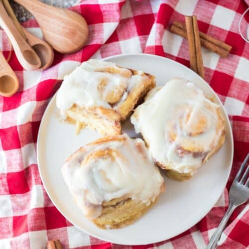 Three cinnamon rolls on a white plate with red and white checkered napkin under the plate.