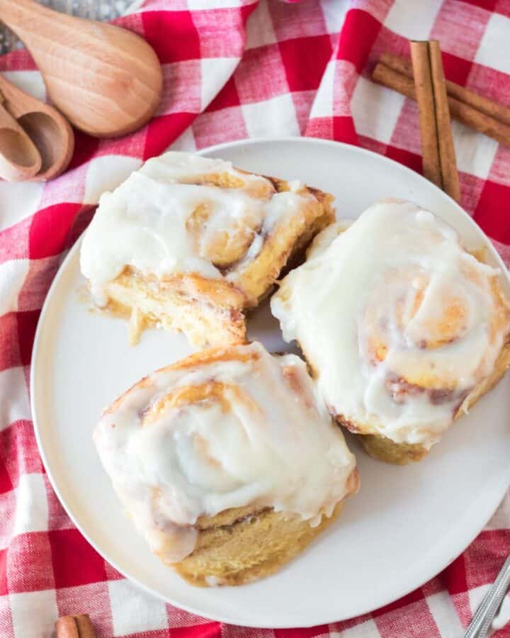 Three cinnamon rolls on a white plate with red and white checkered napkin under the plate.