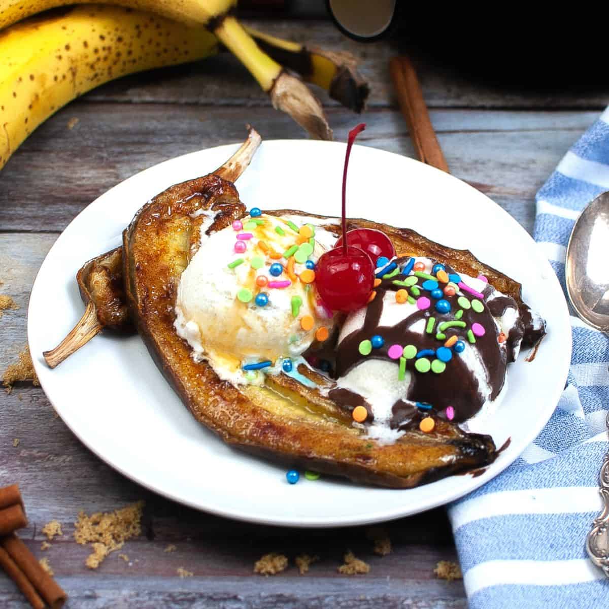 A brown table with a white plate that has bananas that have been air fried and topped with whipped cream, circle sprinkles, and a cherry. 