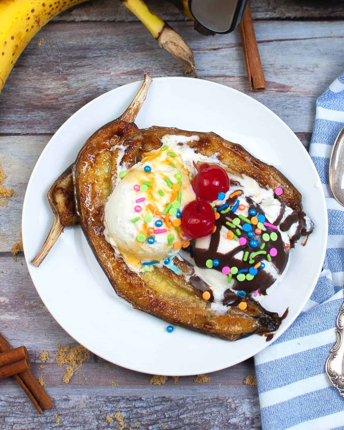 An overhead view of a white plate with bananas that have been air fried topped with ice cream, sprinkles, and  maraschino cherries. 