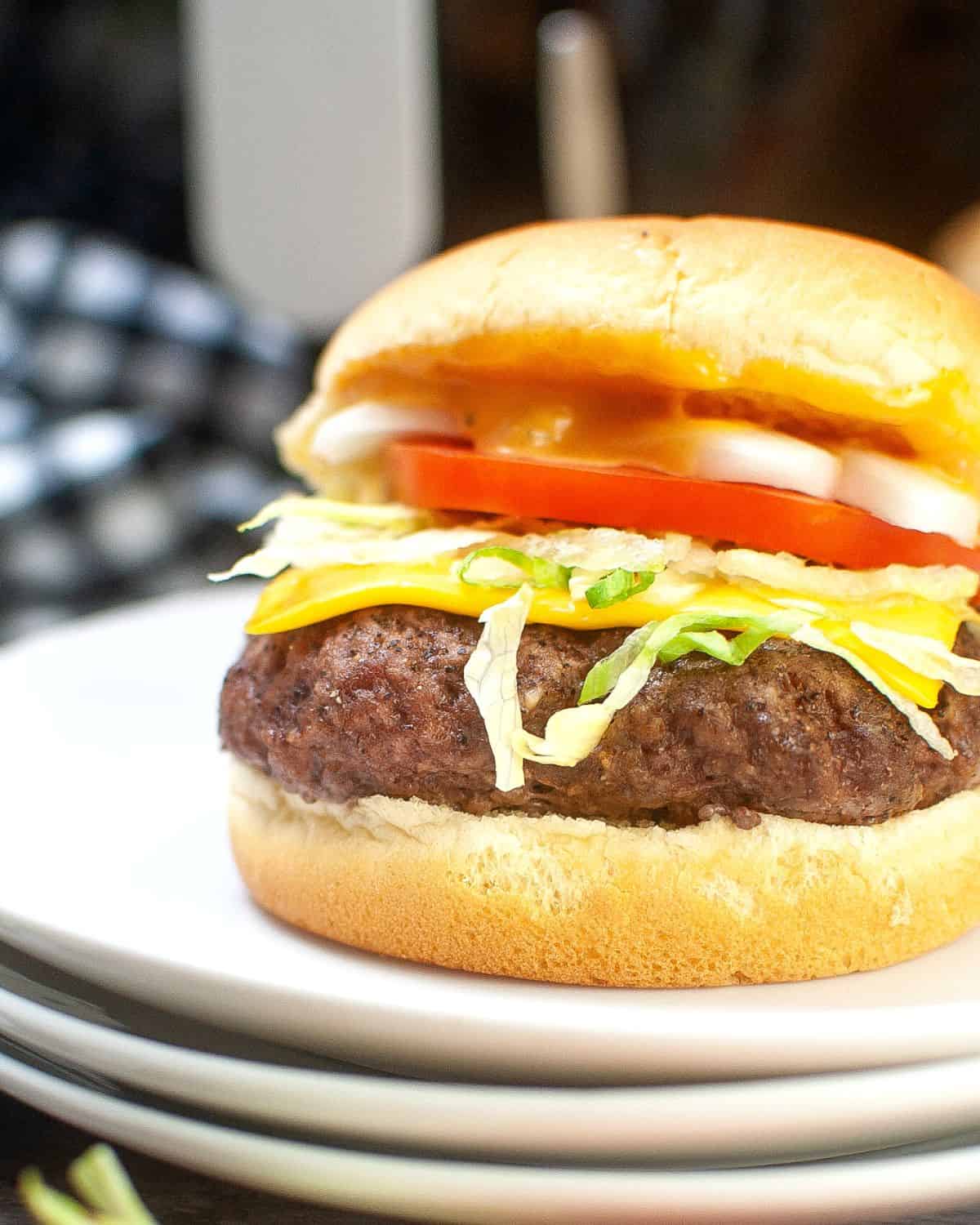 A stack of white plates with a hamburger topped with cheese, tomatoes, and lettuce on the burger. 