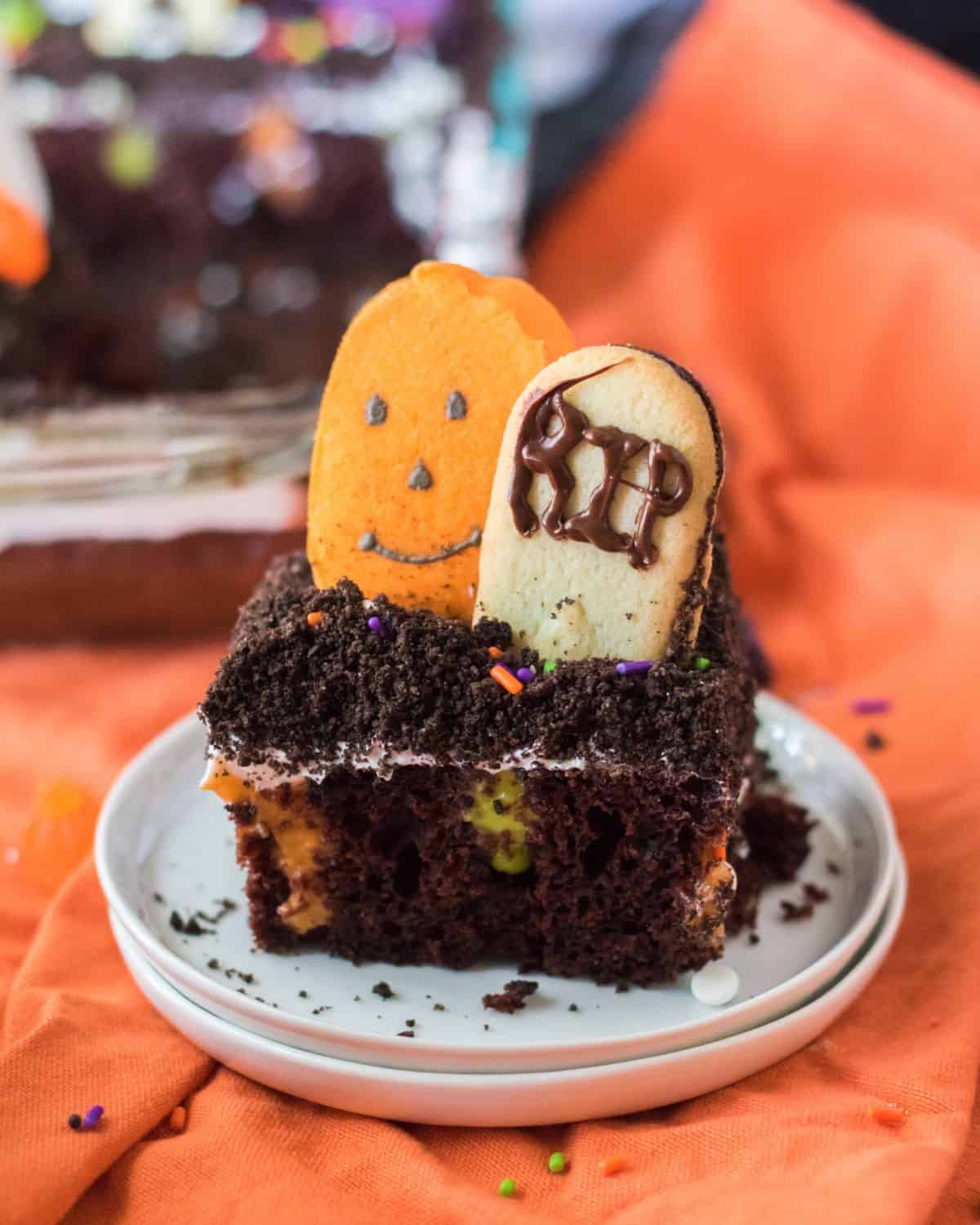 A slice of a chocolate cake decorated fr Haloween with a pumpkin marshmallow on top and a cookie shaped like a tombstone.