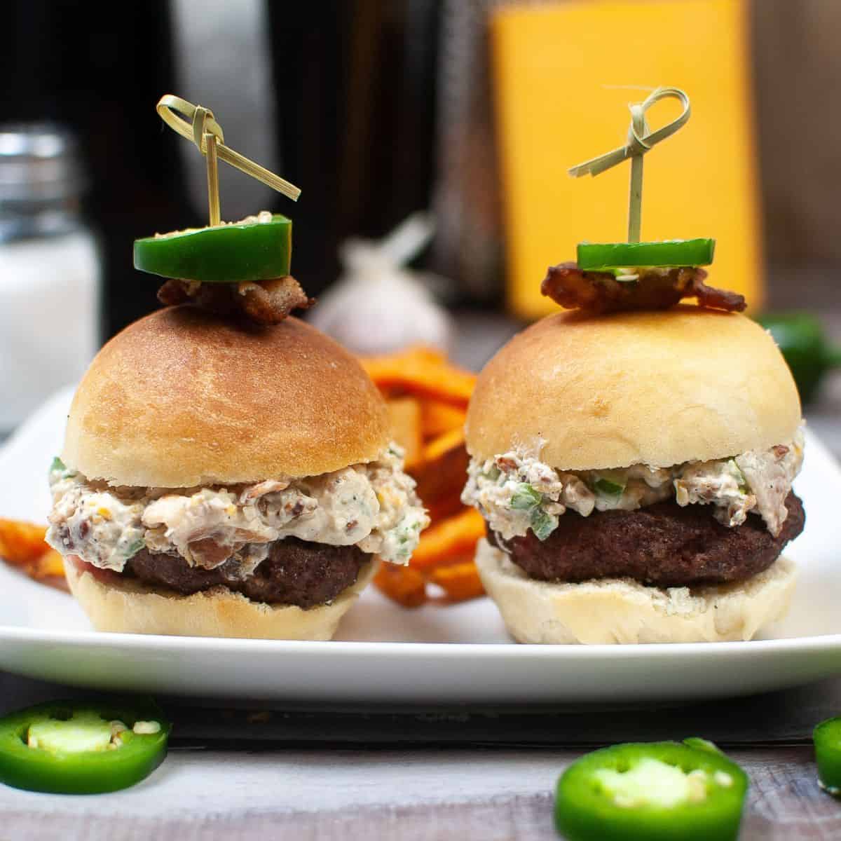 Two hamburger sliders with a creamy topping and the top of the rolls have a slice of bacon and a jalapeno slice on it with a toothpick keeping them in place. 