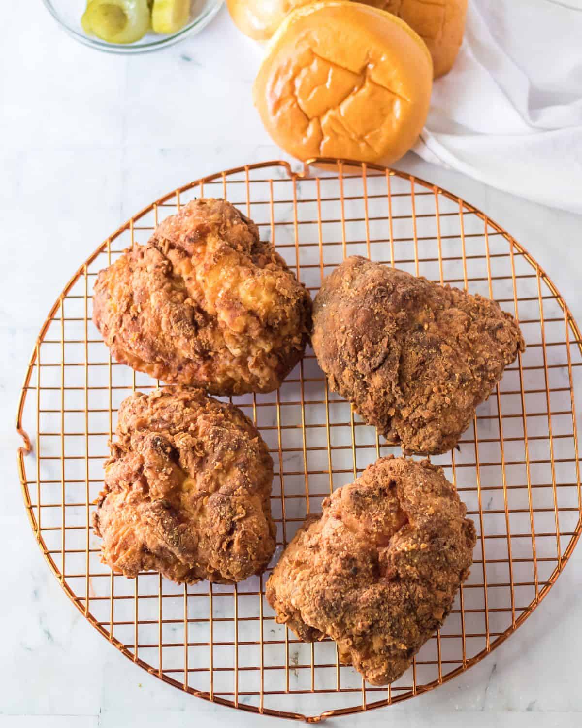 An overhead view of fried chicken breasts on a wire rack with brioche rolls and pickles above the rack to make sandwiches.