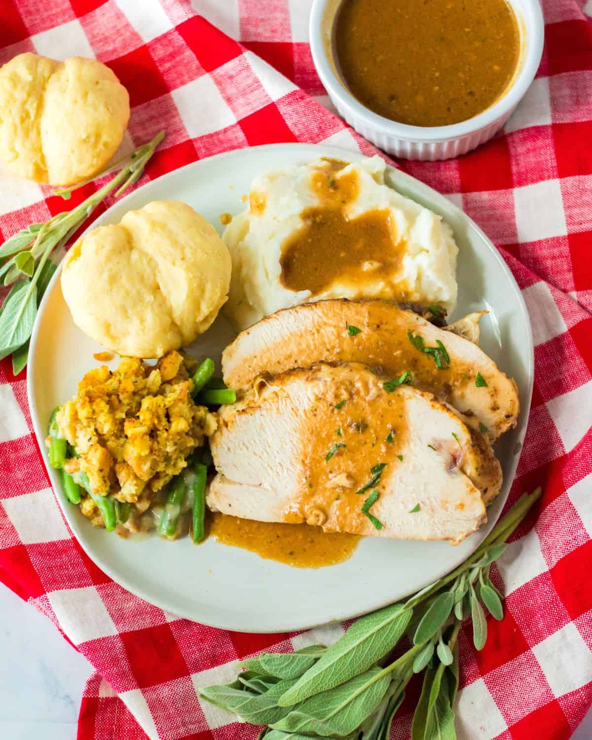 An overhead view of a red and white checkered tablecloth with a plate of turkey mashed potatoes, rolls, and gravy on it.