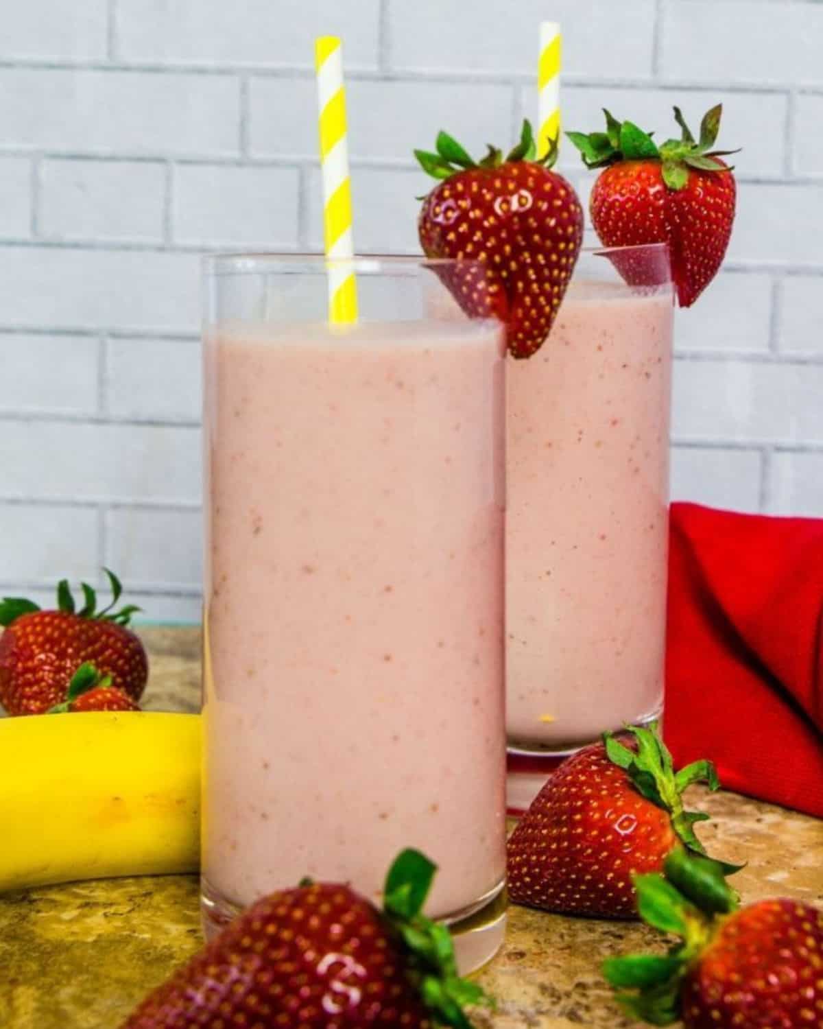 Two tall glasses of fruit smoothie with straws in the glass as well as strawberries on the side of the glass. 