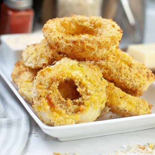 A stack of onion rings on a white plate that are coated in bread crumbs.