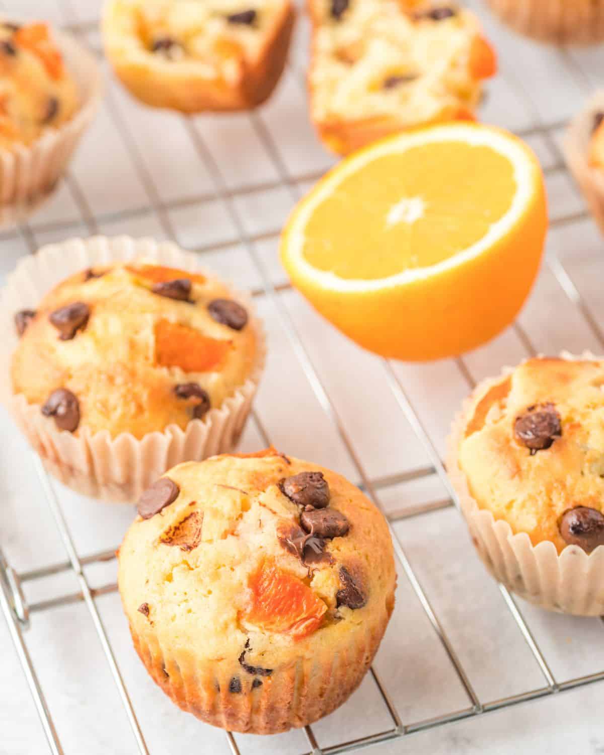 Muffins on a wire rack flavored with pieces of orange and chocolate chips with a half an orange on the side.