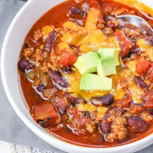 A white bowl filled with beans, cooked ground turkey tomatoes, in a red sauce topped with melted cheddar cheese and diced avocado.