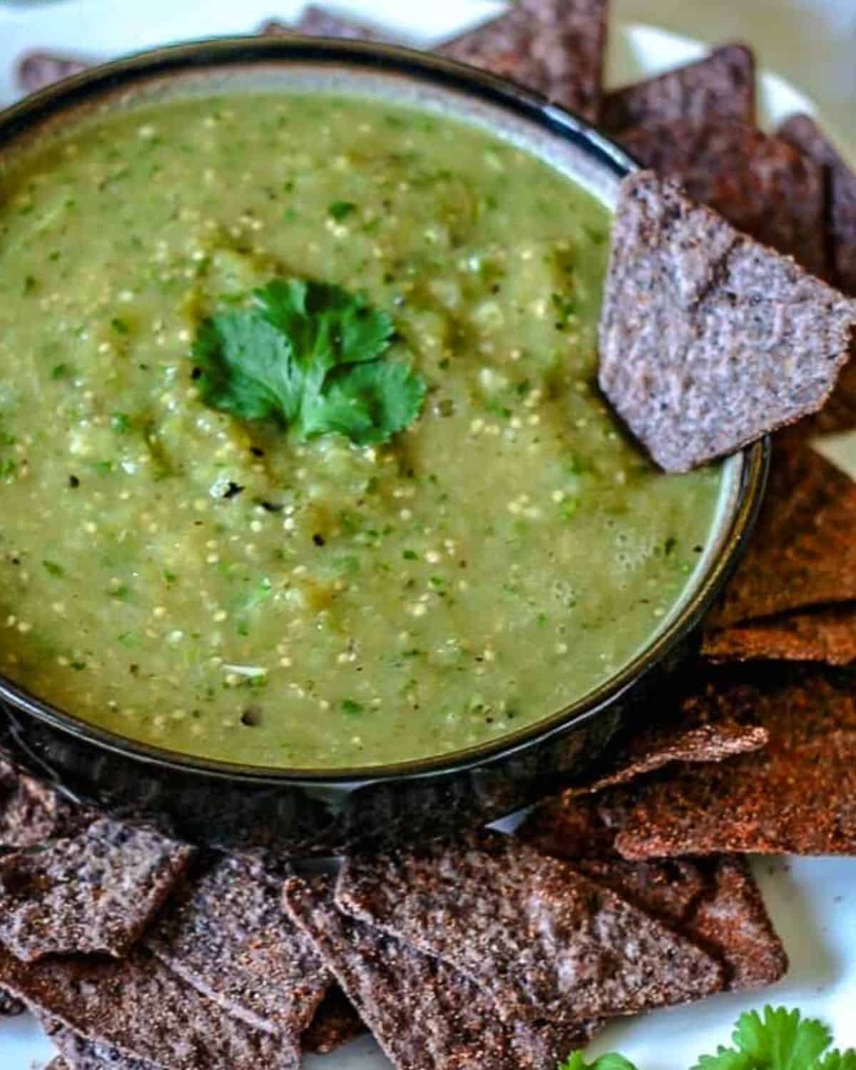 A bowl of green salsa with a blue chip with tortilla chips.