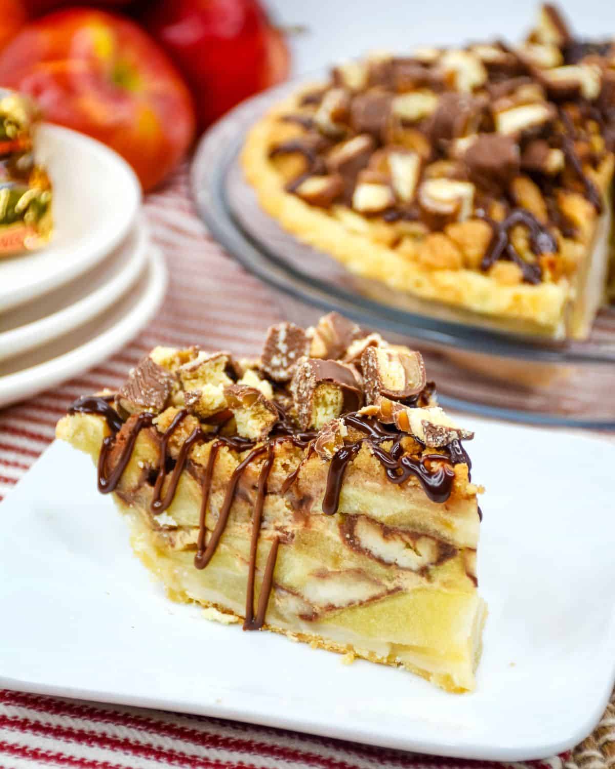 A white plate with a slice of pie stacked with apples and topped with twix candies and a drizzle of chocolate. 