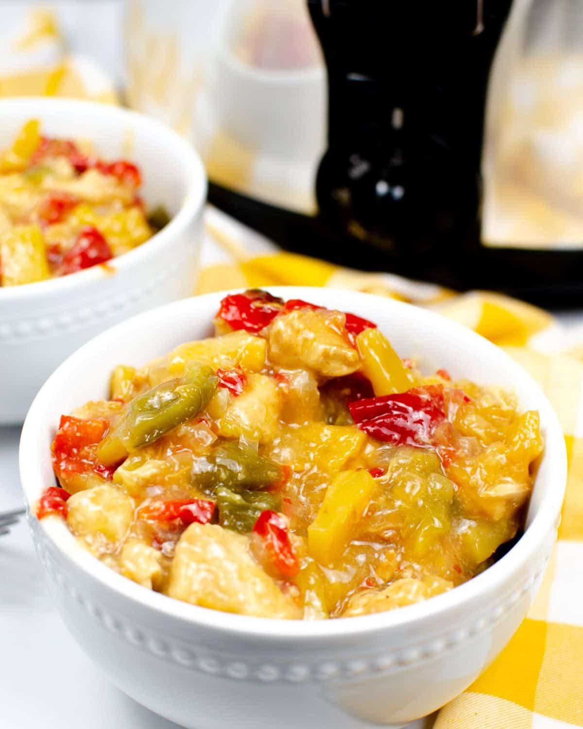 A white bowl on a yellow and white checkerboard linen with an orange sauced meal of chicken and peppers.