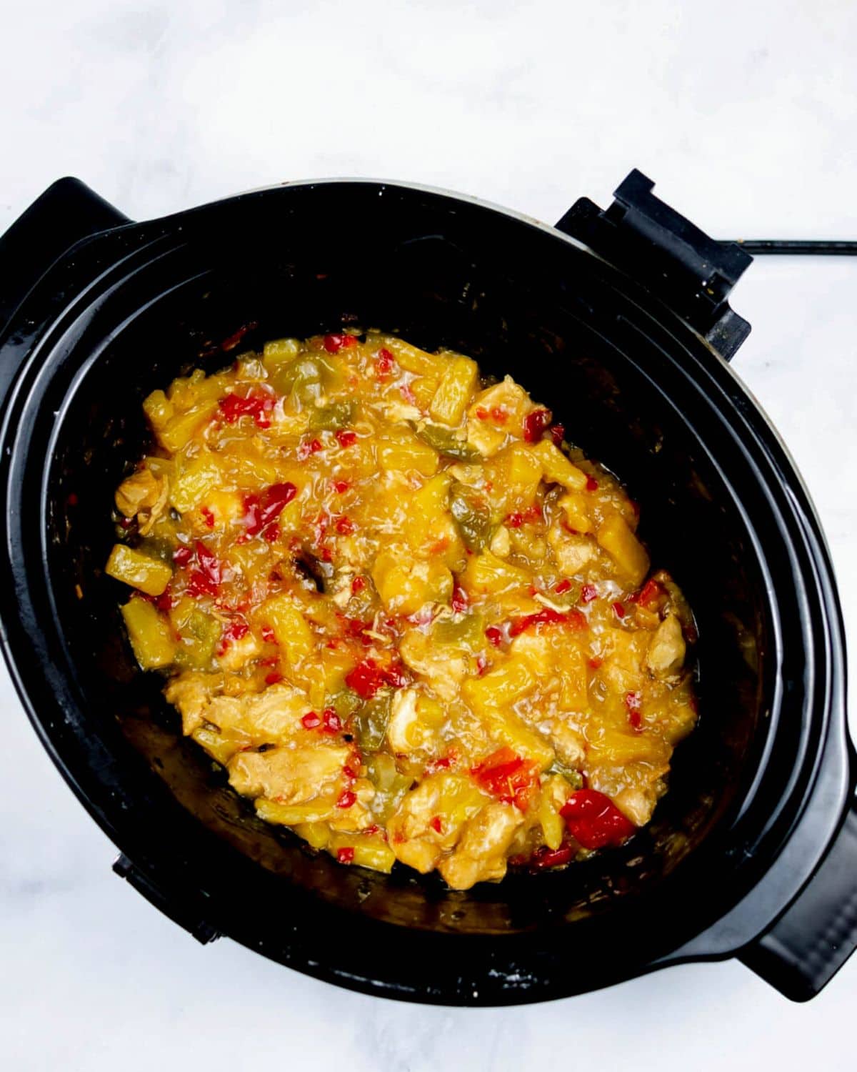 A black slow cooker filled with cooked pineapples, chicken, and peppers in a sauce.