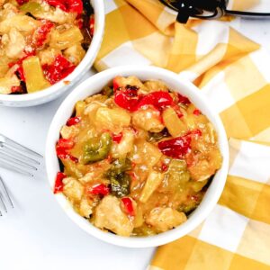 An overhead view of a white bowl on a checkerboard yellow and white linen filled with a chicken and red peppers in a sauce.