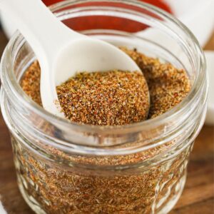 A glass mason jar with seasoning and a spoonful of the seasoning being taken out of the jar.