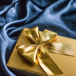 A gold box wrapped with a gold large bow on a blue surface.