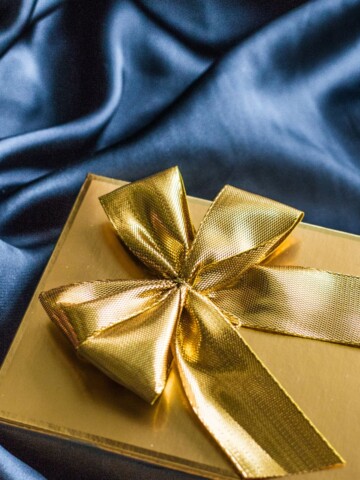 A gold box wrapped with a gold large bow on a blue surface.