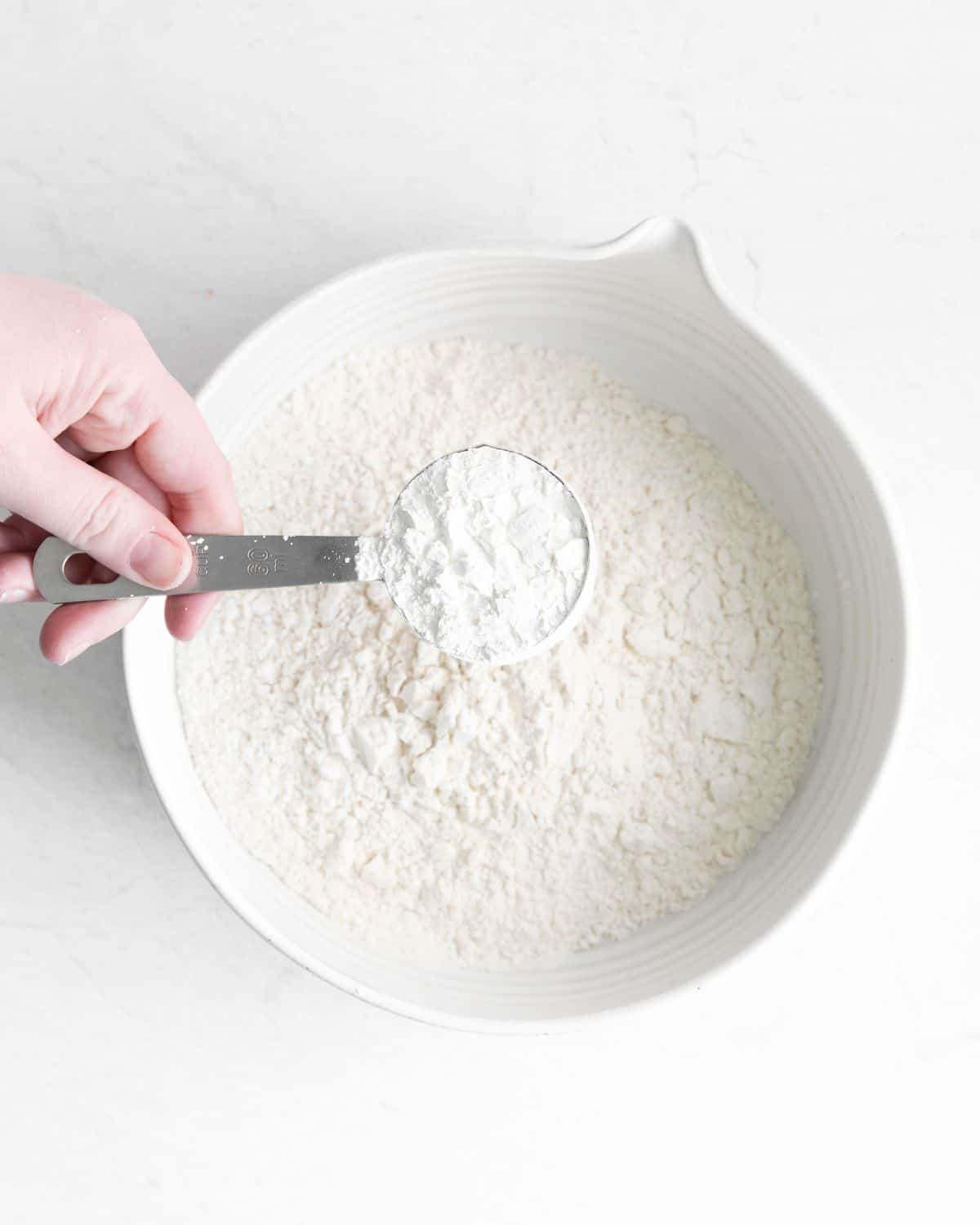 A bowl of all purpose flour with a teaspoon of corn starch being dropped into the bowl. 