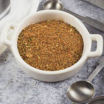 A small white bowl with handles filled with Cajun seasoning with measuring spoon on the side.