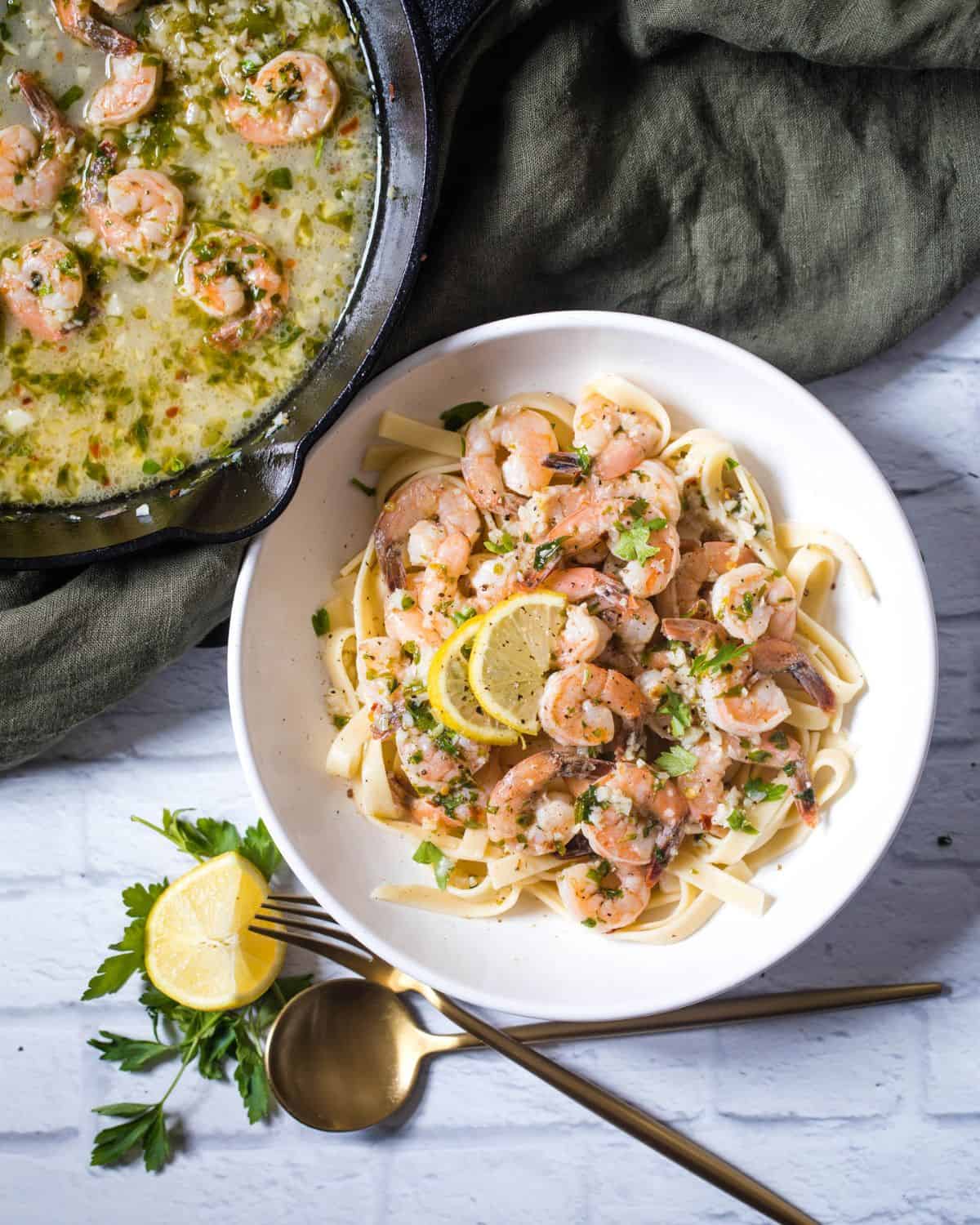 Shrimp Scampi over Linguini on a table with a cast iron pan behind it filled with shrimp in the lemon wine sauce. 