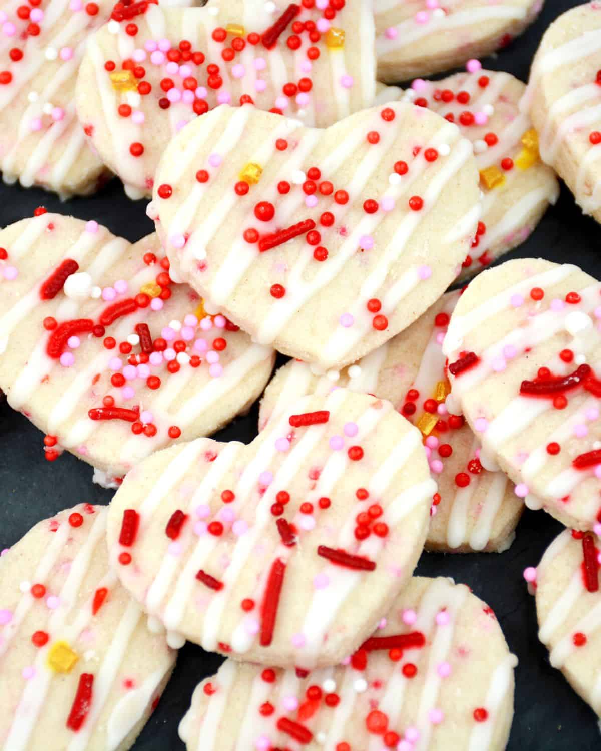 Heart shaped cookies with white frozting drizzle and pink and red sprinkles.