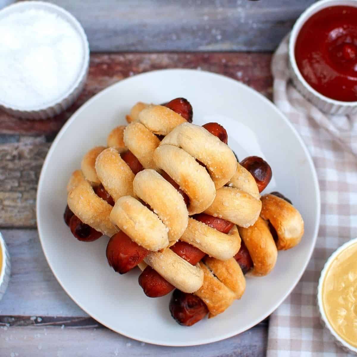Air Fryer Pretzel hot dogs on a plate with dipping sauces on the side.