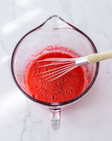 A glass bowl with strawberry gelatin mixed with water using a whisk.