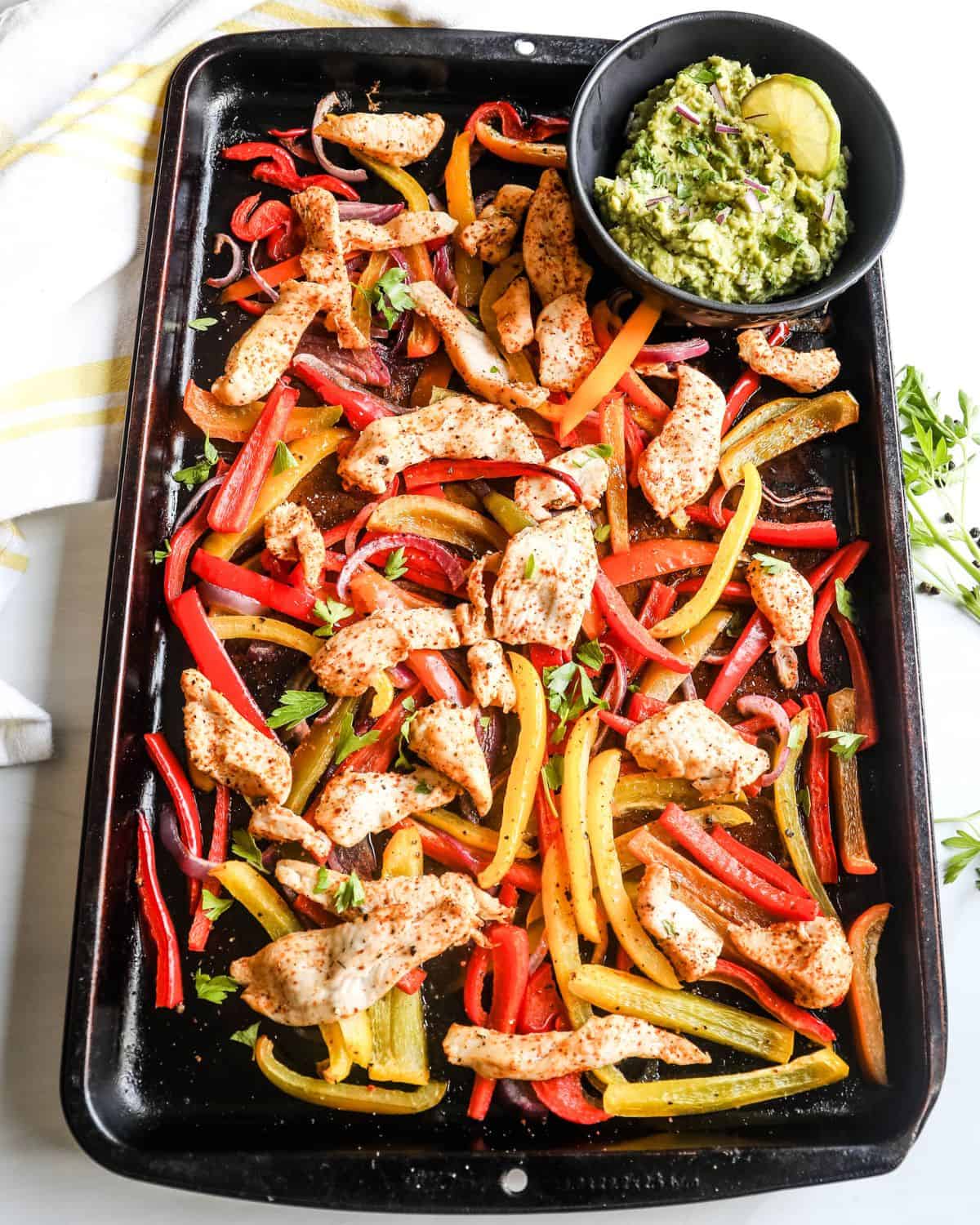 A sheet pan with chicken seasoned with fajita seasoning, sliced peppers, onions and a bowl of guacamole.