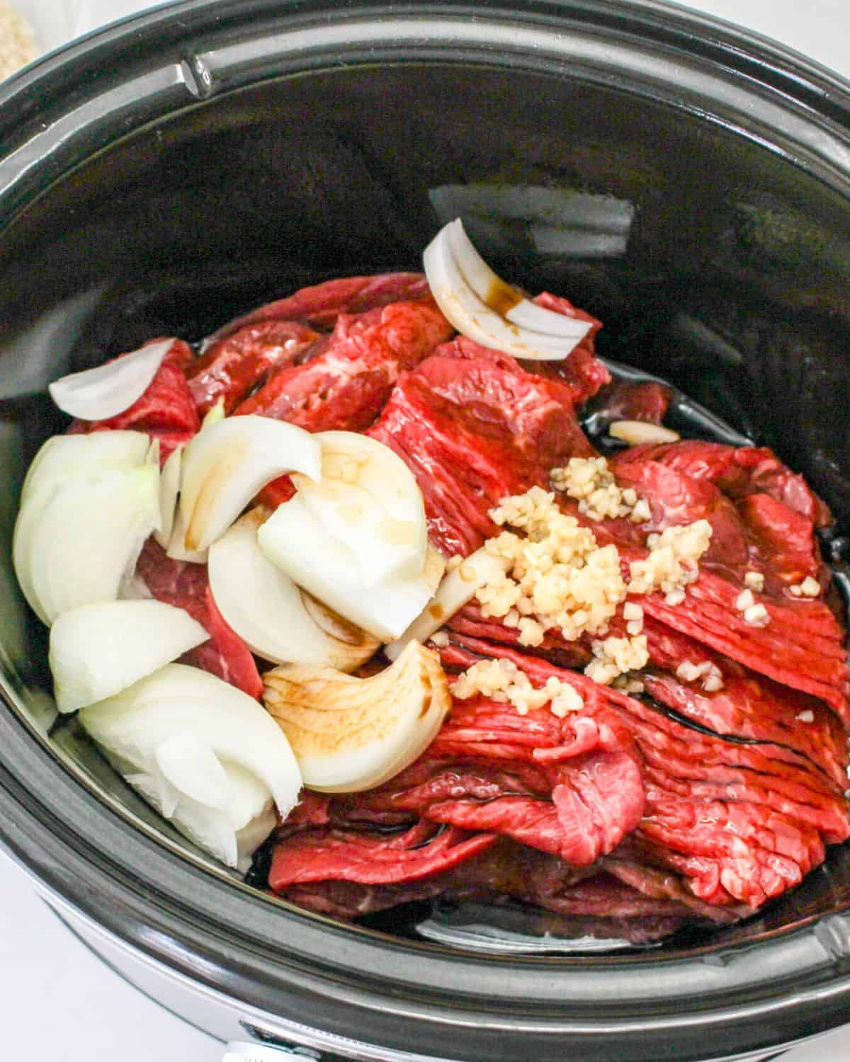 A Slow cooker with beef, onions, garlic, and sauce in the pot.