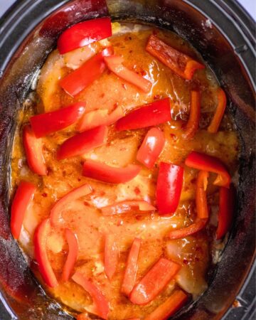 A slow cooker vessel with chicken, chili sauce, and peppers in the vessel. 