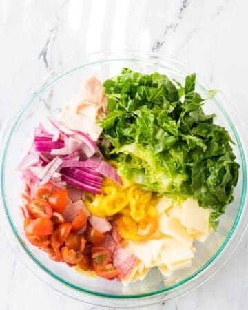 A glass bowl with the step of adding lettuce, red onion, tomatoes, and cold cuts into a bowl to make Italian sub spread.