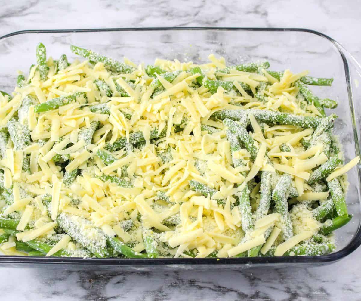 A glass casserole dish with green beans topped with parmesan cheese and gouda cheese.