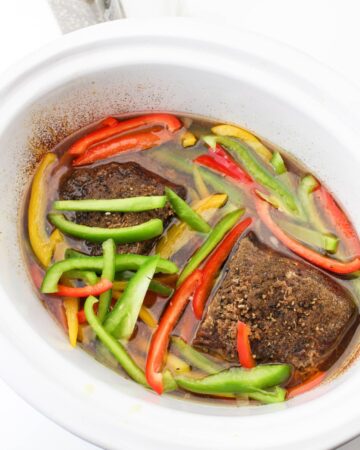 Cooked beef in a slow cooker with sliced peppers being added to the vessel.