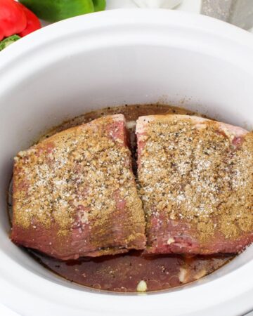 A beef roast seasoned with spiced in a slow cooker vessel to make beef and peppers.