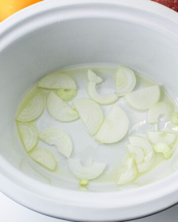 Onions in the bottom of a slow cooker vessel to make beef and peppers.