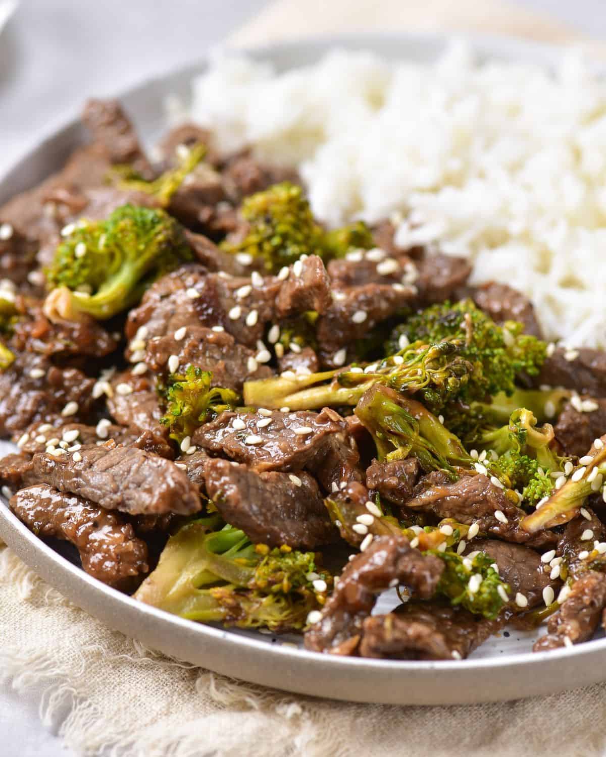 A white plate with rice on one side and beef and broccoli topped with sesame seeds on the left side of the plate.