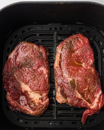 An air fryer basket with seasoned boneless rib eye steaks ready to be air fried.